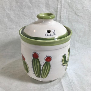 Può includere: Barattolo di ceramica bianco con coperchio verde e disegni di cactus. Il coperchio ha un foro e la parola "olio" scritta su di esso.