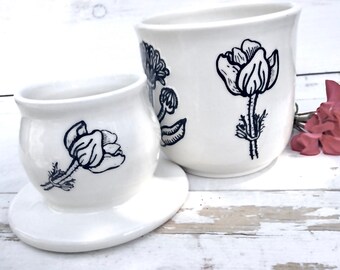 Könnte beinhalten: Zwei weiße Keramik-Butterdosen mit schwarzen Blumendesigns. Die kleinere Dose hat einen Deckel und eine Untertasse. Die größere Dose ist höher und hat ein einzelnes Blumendesign.