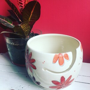 Puede incluir: Cuenco de lana de cer&aacute;mica blanca con un dise&ntilde;o floral rojo y naranja. El cuenco tiene un agujero en el lateral para que la lana pueda pasar. Un gancho de ganchillo de madera est&aacute; en primer plano.