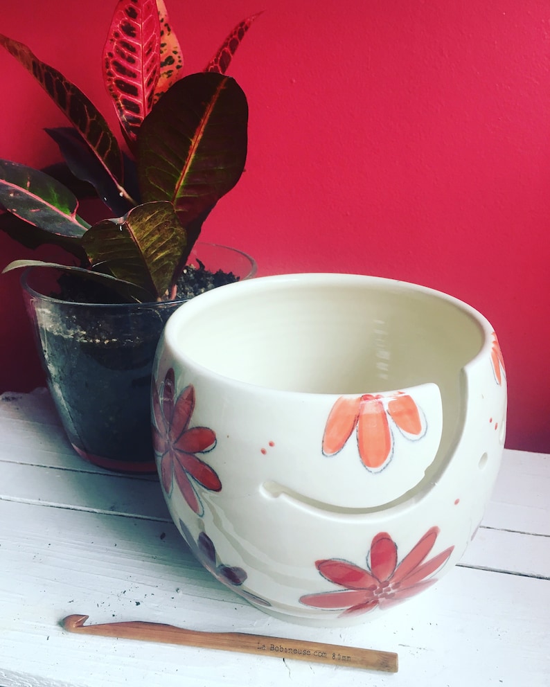 Puede incluir: Cuenco de lana de cer&aacute;mica blanca con un dise&ntilde;o floral rojo y naranja. El cuenco tiene un agujero en el lateral para que la lana pueda pasar. Un gancho de ganchillo de madera est&aacute; en primer plano.