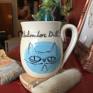 May include: White ceramic mug with a blue cat illustration and the text "Salvador Dali" in black script.