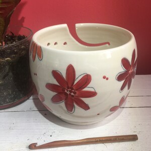 Puede incluir: Cuenco de lana de cer&aacute;mica blanca con un dise&ntilde;o floral rojo y blanco. El cuenco tiene un agujero en el lateral para que la lana pueda pasar. Un gancho de ganchillo de madera est&aacute; en primer plano.