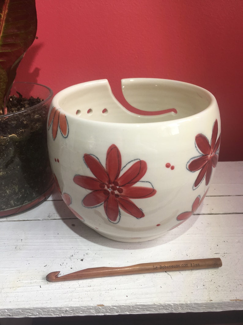 Puede incluir: Cuenco de lana de cer&aacute;mica blanca con un dise&ntilde;o floral rojo y blanco. El cuenco tiene un agujero en el lateral para que la lana pueda pasar. Un gancho de ganchillo de madera est&aacute; en primer plano.