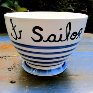 Op de afbeelding: Een witte keramische kom met blauwe en witte strepen en de woorden "Le Sailor" in het zwart. De kom heeft een blauwe en witte voet.