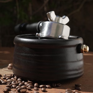 May include: A black ceramic espresso machine portafilter holder with a silver portafilter. Coffee beans are scattered around the base. The background is blurred, with a natural, earthy tone.