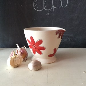 Può includere: Un'anfora in ceramica bianca con un motivo floreale rosso. L'anfora ha un beccuccio e un manico. Ci sono tre spicchi d'aglio sul tavolo accanto all'anfora.