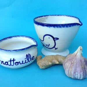 Può includere: Due ciotole in ceramica bianca con bordo blu. La ciotola più piccola ha la scritta "Grattouille". La ciotola più grande ha un disegno di un uccello blu. Un pezzo di zenzero e uno spicchio d'aglio viola sono davanti alle ciotole.