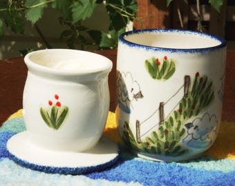 May include: Two white ceramic pieces with blue trim. One is a small container with a lid, decorated with a green and red floral design. The other is a tall mug with a sheep and fence design. Both are on a colorful striped surface.