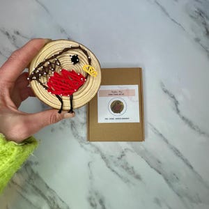 May include: A wooden round ornament with a red robin bird design. The bird is made with string art and has a yellow beak and black eyes. The ornament is packaged in a brown box with the text "Make Me Robin String Art Kit" and "Pine String Wooden Ornament".