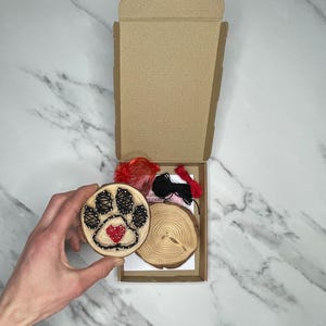 May include: A DIY string art kit featuring a wooden circle with a black paw print and a red heart in the centre. The kit includes red and black string, a wooden circle, and a small wooden heart.