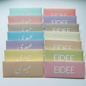 May include: A collection of pastel-colored rectangular envelopes. Each envelope features cut-out text, with some displaying the word "EIDEE" and others displaying Arabic script. The envelopes are arranged in neat rows, showcasing a variety of soft hues.