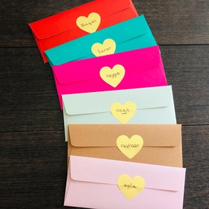 May include: A stack of colorful envelopes in red, teal, pink, light green, tan, and light pink. Each envelope has a yellow heart-shaped sticker with a name written on it. The names are Khaqan, Faran, Rayya, Noah, Rayhaan, and Ayla.
