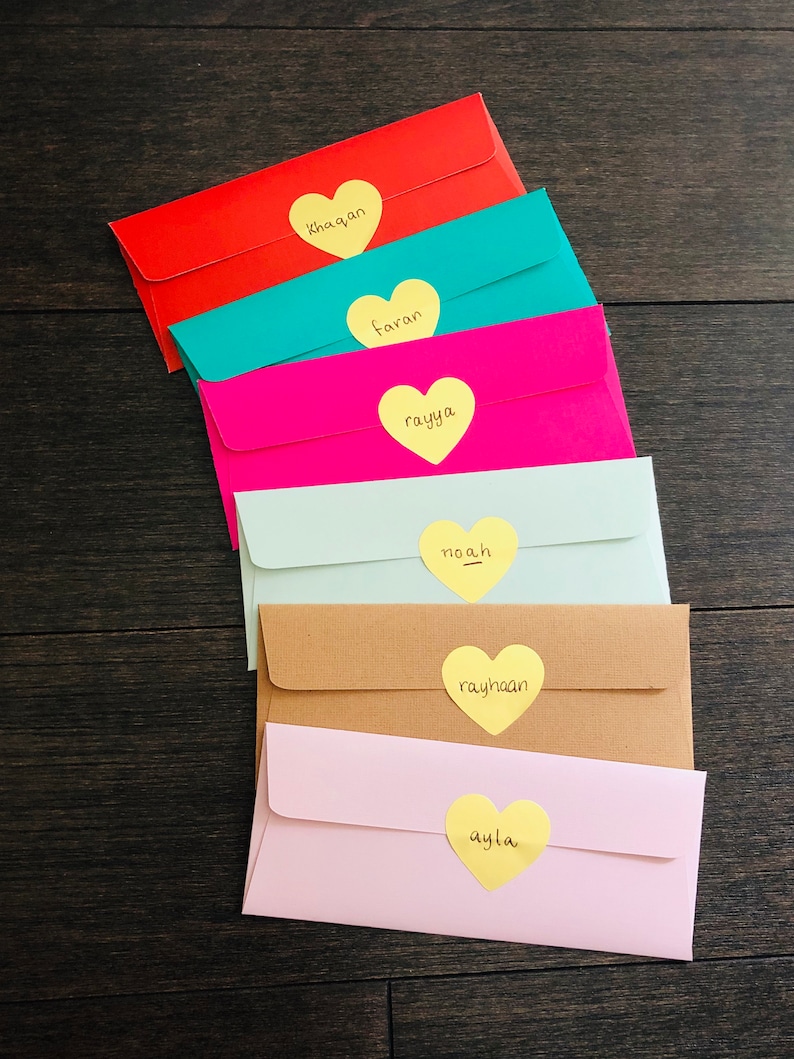 May include: A stack of colorful envelopes in red, teal, pink, light green, tan, and light pink. Each envelope has a yellow heart-shaped sticker with a name written on it. The names are Khaqan, Faran, Rayya, Noah, Rayhaan, and Ayla.