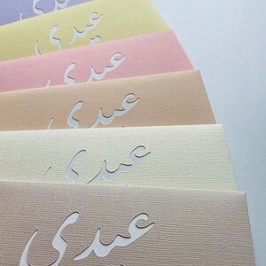 May include: A collection of pastel-colored cards, each featuring a cutout Arabic script. The cards are arranged in a fan shape, showcasing a variety of soft hues including lavender, yellow, pink, and beige. The paper has a textured finish.
