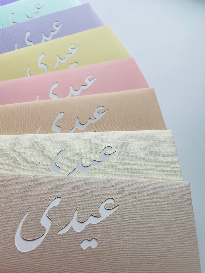 May include: A collection of pastel-colored cards, each featuring a cutout Arabic script. The cards are arranged in a fan shape, showcasing a variety of soft hues including lavender, yellow, pink, and beige. The paper has a textured finish.