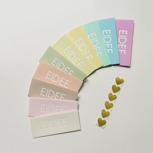 May include: A collection of pastel-colored rectangular cards arranged in a semi-circle, each bearing the embossed word "EIDEE" in white. A strip of gold heart-shaped stickers is positioned to the right, adding a decorative element to the composition.