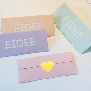 May include: Four pastel-colored envelopes. Three rectangular envelopes are peach, lavender, and mint green, each with the word "EIDEE" in white. A fourth, pink envelope has a yellow heart sticker.