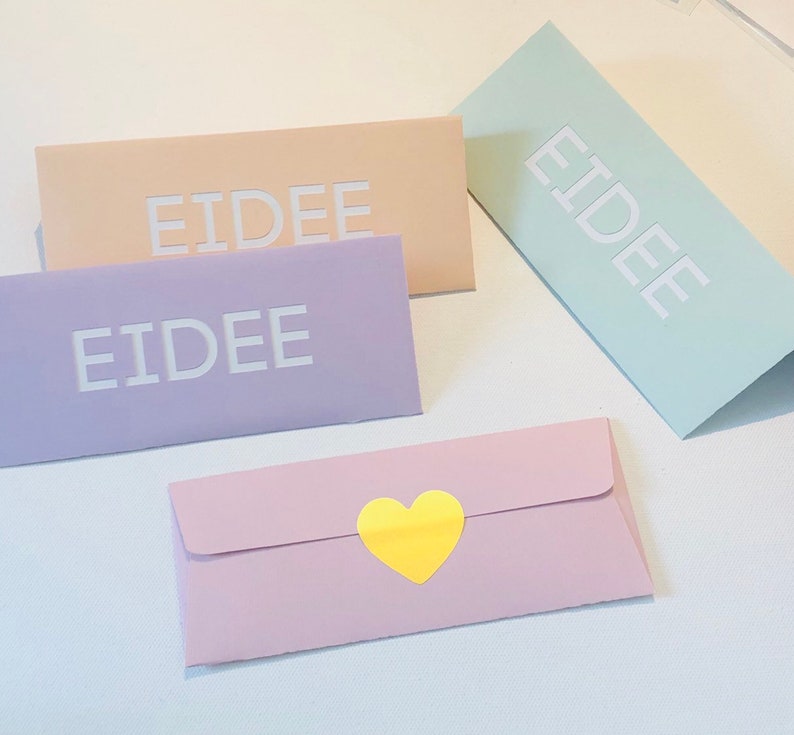 May include: Four pastel-colored envelopes. Three rectangular envelopes are peach, lavender, and mint green, each with the word "EIDEE" in white. A fourth, pink envelope has a yellow heart sticker.