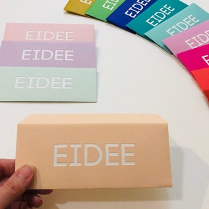 May include: A collection of pastel-colored envelopes with the word "EIDEE" embossed in white. The envelopes are in shades of pink, lavender, mint green, and peach. A hand holds a peach-colored envelope with the same text.
