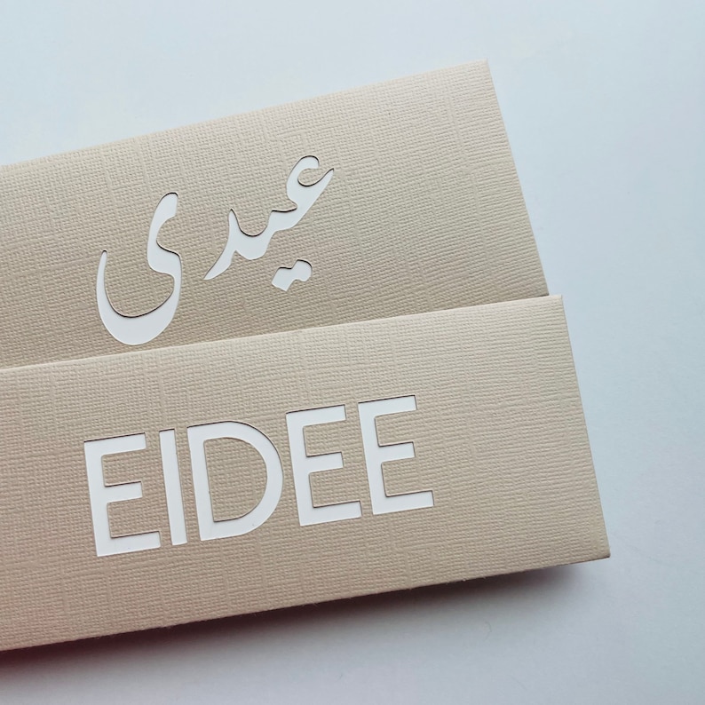 May include: Beige cardstock with the word "EIDEE" cut out in white block letters. Above, the Arabic word "عيدي" is also cut out. The cardstock has a textured surface and is folded.