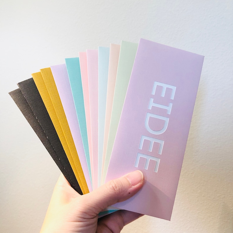 May include: A hand holds a fan of pastel-colored envelopes. The envelopes are in shades of black, gold, light blue, pink, and lavender. The word "EIDEE" is embossed in white on the lavender envelope.