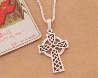 May include: A silver-colored Celtic cross pendant with an intricate openwork design, suspended from a delicate chain. The cross features a circular halo and a detailed knot pattern. A vintage postcard with French text is in the background.