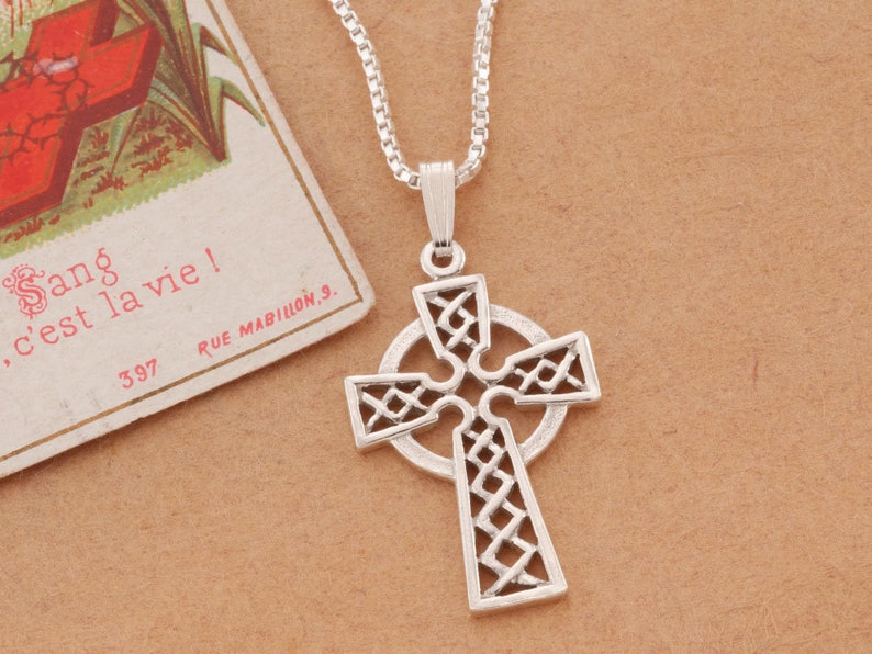 Peut inclure: Pendentif croix celtique argent&eacute; avec un motif ajour&eacute; complexe, suspendu &agrave; une cha&icirc;ne d&eacute;licate. La croix pr&eacute;sente un halo circulaire et un motif de n&oelig;ud d&eacute;taill&eacute;. Une carte postale vintage avec du texte fran&ccedil;ais est en arri&egrave;re-plan.