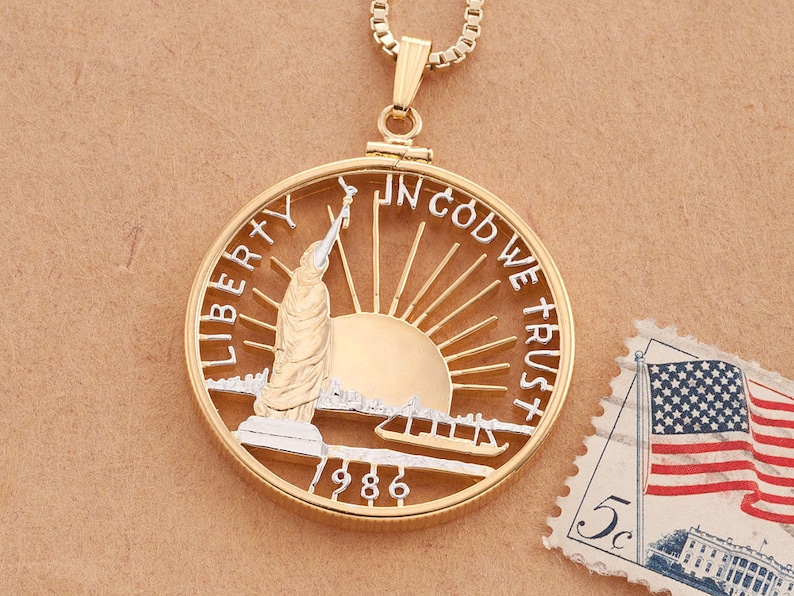 May include: Gold-tone pendant necklace featuring a cutout design of the Statue of Liberty, a sun, and the words "Liberty", "In God We Trust", and "1986".