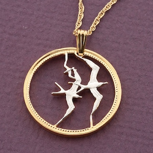 May include: A gold pendant necklace with a circular design. The pendant features two silver-toned birds in flight within a gold frame, suspended from a delicate gold chain. The background is a deep purple.