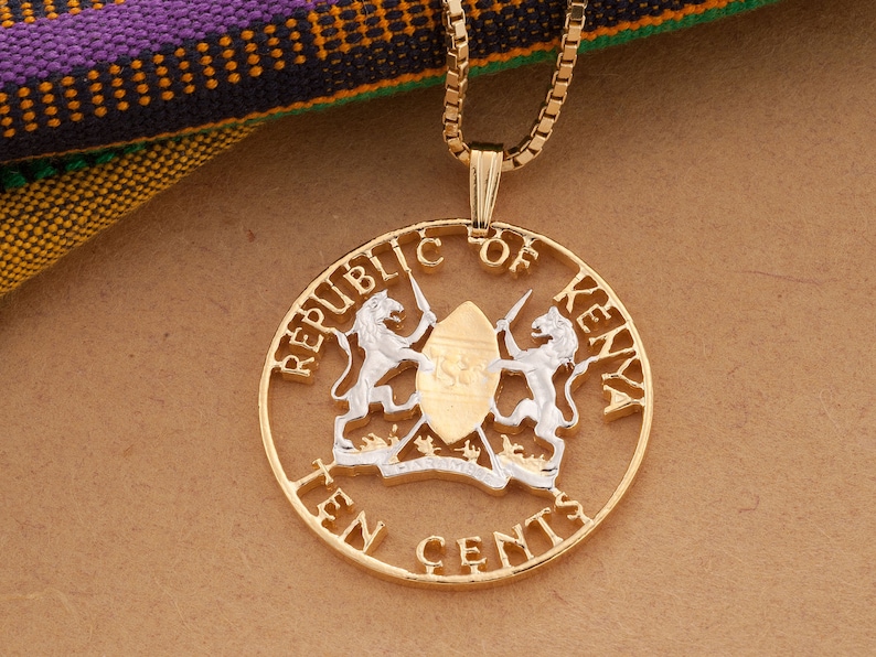 May include: Gold-coloured pendant necklace featuring the emblem of the Republic of Kenya. The pendant has a circular design with the words "REPUBLIC OF KENYA" and "TEN CENTS" around the edge. The centre features a shield and two lions. The necklace chain is also gold-coloured.