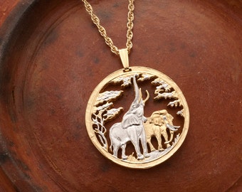 Puede incluir: Collar con colgante dorado con un diseño circular que representa dos elefantes y árboles. Los elefantes son en tonos plateados y dorados. El colgante cuelga de una cadena dorada. El collar se muestra sobre una superficie marrón.