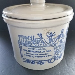 May include: White ceramic crock with a blue and white design. The design features a hobby horse, drums, trumpets, and the text "Hobby-Horses, Pop Settlers and Dolls, Dancing Jacks, Drums, Trumpets."