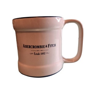 May include: A white ceramic mug with a black rim and handle. The mug has the text "Abercrombie & Fitch" and "Estab. 1892" printed on the front.