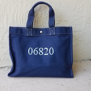 May include: A navy blue tote bag with white stitching and handles. The bag features the number "06820" printed in white on the front. The tote bag is made of canvas material and has a button closure.