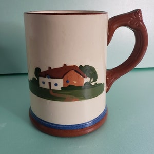 May include: A white ceramic mug with a brown handle. The mug has a painted scene of a white house with a brown roof and green trees. The house has blue windows and a blue door. The mug has a blue and brown band around the rim.