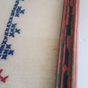 May include: A close-up of a framed piece of needlepoint embroidery. The embroidery features a blue and red design on a white background. The frame is made of wood and has a red, blue, and green paint finish.