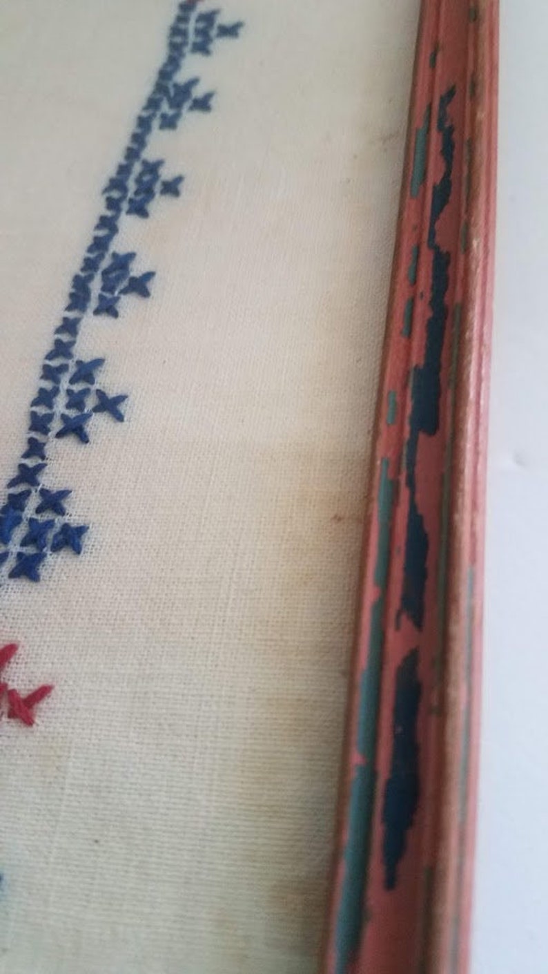 May include: A close-up of a framed piece of needlepoint embroidery. The embroidery features a blue and red design on a white background. The frame is made of wood and has a red, blue, and green paint finish.