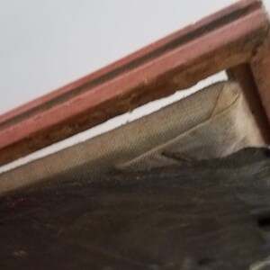 May include: Close-up of the top edge of a framed picture. The frame is made of wood and has a red-brown finish. The picture is covered with a layer of fabric.