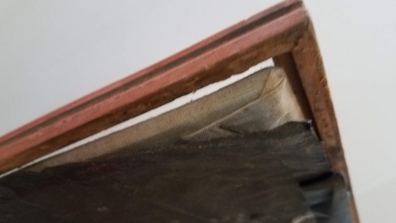 May include: Close-up of the top edge of a framed picture. The frame is made of wood and has a red-brown finish. The picture is covered with a layer of fabric.