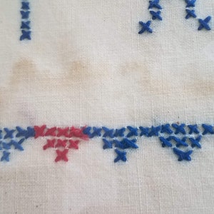May include: Close-up of a white fabric with blue and red cross-stitch embroidery. The embroidery forms a border along the bottom of the fabric.