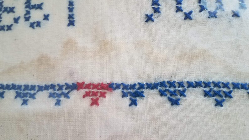 May include: Close-up of a white fabric with blue and red cross-stitch embroidery. The embroidery forms a border along the bottom of the fabric.