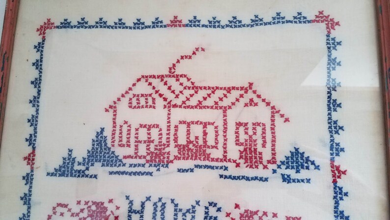 May include: A framed cross-stitch embroidery depicting a red house with a blue chimney and two blue trees. The text "Home Sweet Home" is stitched below the house.