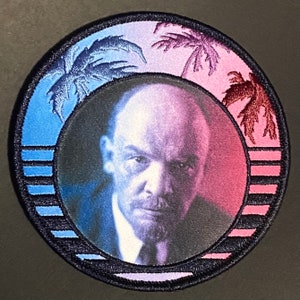 May include: A round embroidered patch with a portrait of a man in the center. The background is a gradient of blue and pink with palm trees on either side. The patch has a black border and a white stripe pattern around the edge.