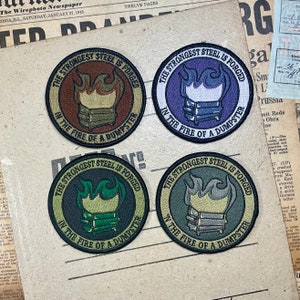 Pode incluir: Quatro patches bordados circulares com o texto "THE STRONGEST STEEL IS FORGED IN THE FIRE OF A DUMPSTER". Cada patch apresenta um caixote do lixo e chamas em diferentes esquemas de cores: castanho, roxo, verde e cinzento. Os patches estão num fundo de jornal vintage.