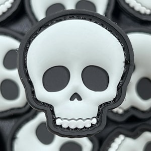 May include: A close-up of a white skull patch with black eyes and a black outline. The patch is made of a soft, flexible material and has a velcro backing.
