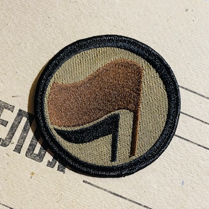 Könnte beinhalten: Ein braunes und schwarzes gesticktes Patch mit einem stilisierten Flaggen-Design. Die Flagge ist braun mit einem schwarzen Griff und befindet sich auf einem grünen Hintergrund. Das Patch ist von einem schwarzen Rand umgeben.