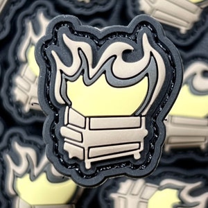 May include: A black, tan, and yellow shoe charm featuring a stylized couch engulfed in flames. The charm has a black border and a textured appearance, with the couch and flames in contrasting colors.