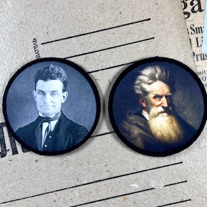 Op de afbeelding: Twee ronde zwarte en witte patches met portretten van mannen. De patch links toont een man met kort donker haar, een witte kraag en een donkere stropdas. De patch rechts toont een man met lang wit haar en een witte baard.