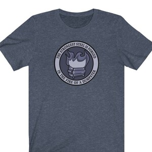 Pode incluir: Uma camiseta azul escura com um design gráfico circular. O gráfico apresenta um caixote do lixo em chamas com o texto "The strongest steel is forged in the fire of a dumpster".