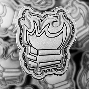 May include: A silver-coloured pin featuring a design of a stack of books with flames above. The pin has a detailed outline and a textured border, creating a three-dimensional effect. Other similar pins are visible in the background.